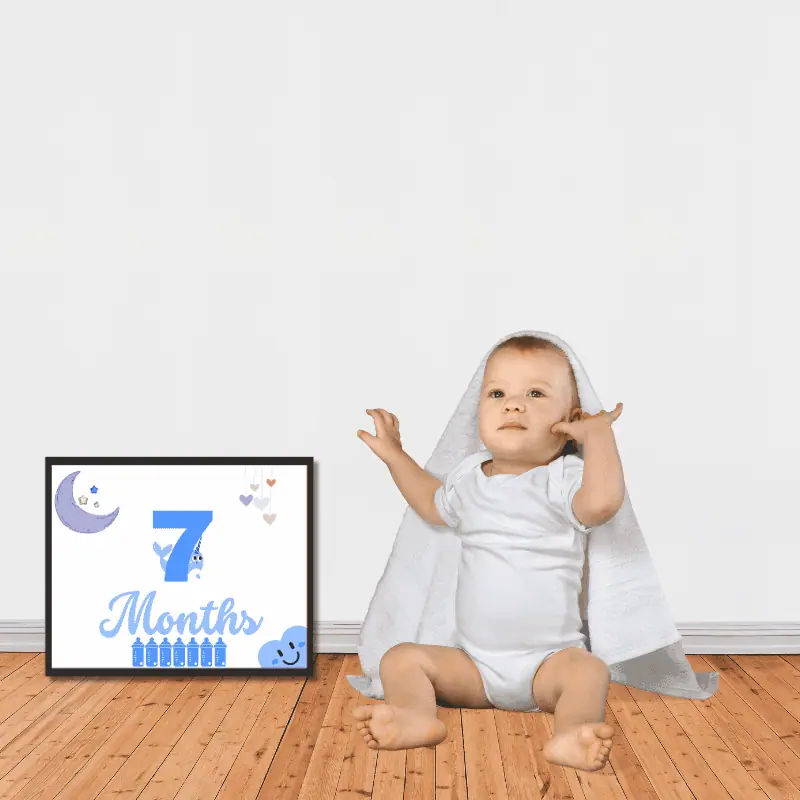 A milestone sign in complimentary shades of blue framed next to a baby sitting on the floor. The sign says 7 months and features cute, baby themed decoration.