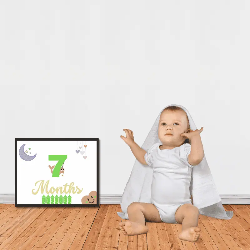A milestone sign in shades of green and gold framed next to a baby sitting on the floor. The sign says 7 months and features cute, baby themed decoration.