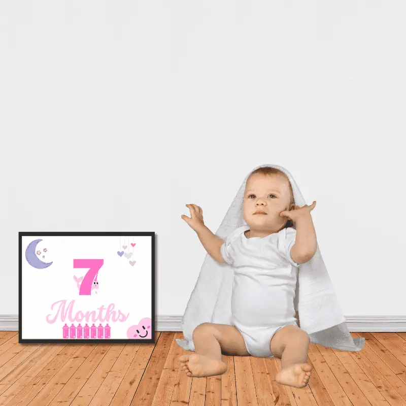 A milestone sign in complimentary shades of pink framed next to a baby sitting on the floor. The sign says 7 months and features cute, baby themed decoration.