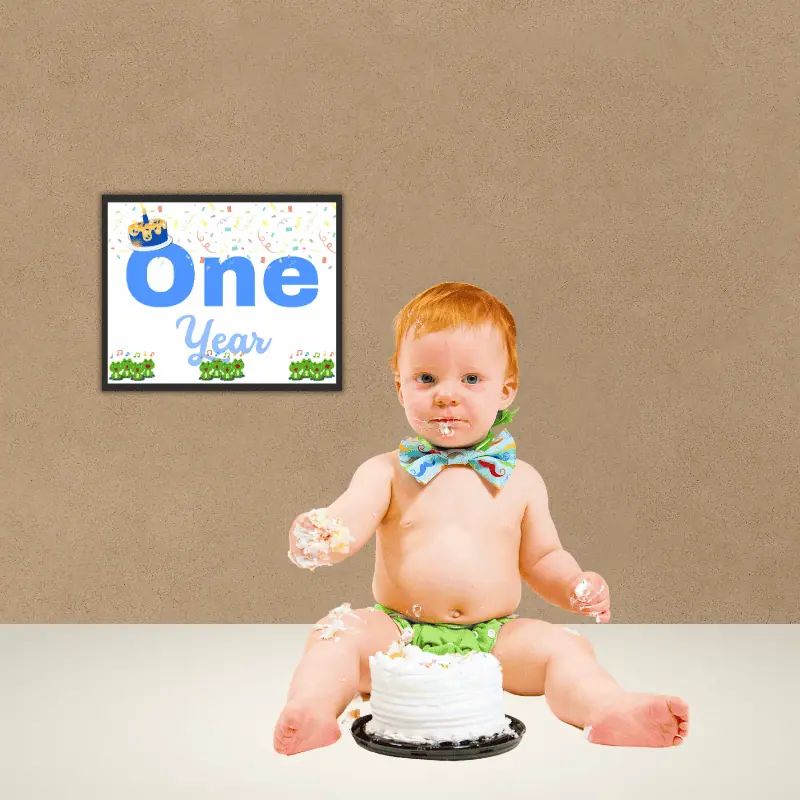 A baby milestone sign that says one year in complimentary shades of blue and features 12 singing frogs, a cake with one candle and confetti framed on the wall next to a baby boy eating a small birthday cake very messily.
