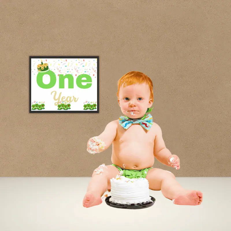 A baby milestone sign that says one year in complimentary shades of teal and features 12 singing frogs, a cake with one candle and confetti framed on a wall beside a baby sitting on the floor eating cake.