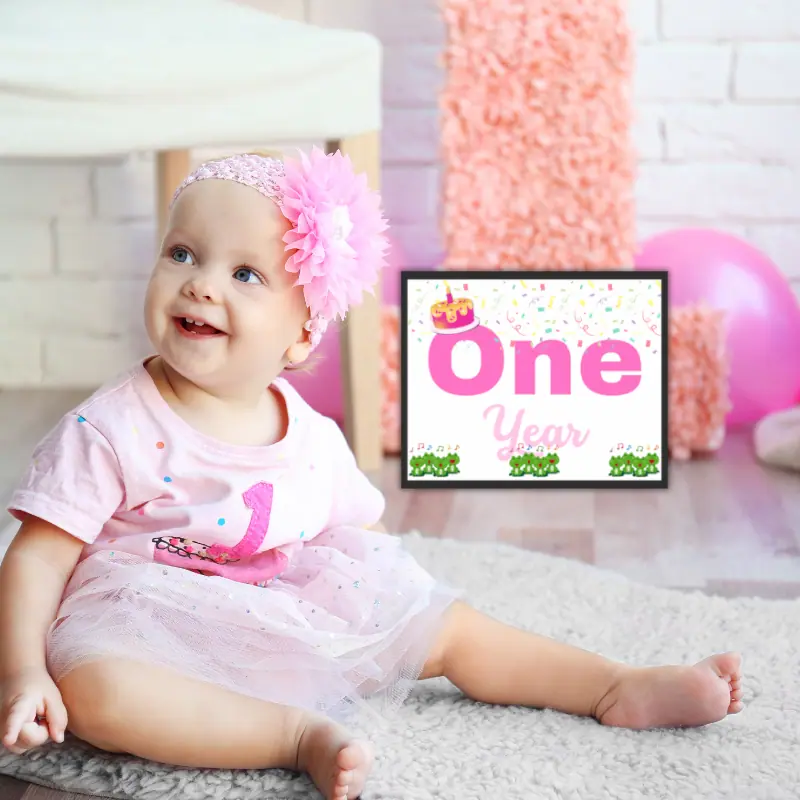 A baby milestone sign that says one year in complimentary shades of pink and features 12 singing frogs, a cake with one candle and confetti framed leaned up against birthday decorations next to a baby girl in pink smiling.