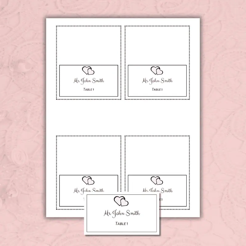 a folded and a flat wedding place card printable with a customizable thin border and 2 hearts laying on a table
