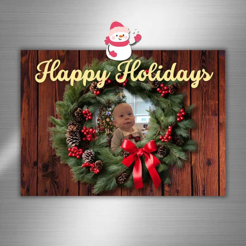 A holiday photo card has photo of a baby peaking out from the middle of the wreath hanging on the fridge.