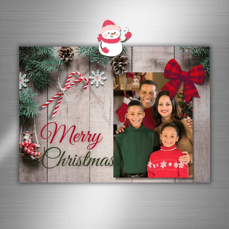 A Christmas Photo Card hanging on the fridge. The card has winter greens, pine cones, and a candy cane around a photo with a fluffy red bow in the corner.